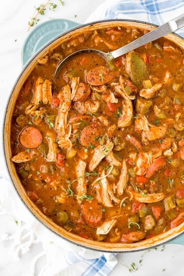 A pot of gumbo sits on a white countertop and covers a large portion of the image. In the gumbo are several ingredients, including chicken, sausage, okra, tomatoes, and various herbs as a garnish. There is a ladle in the gumbo that is full.