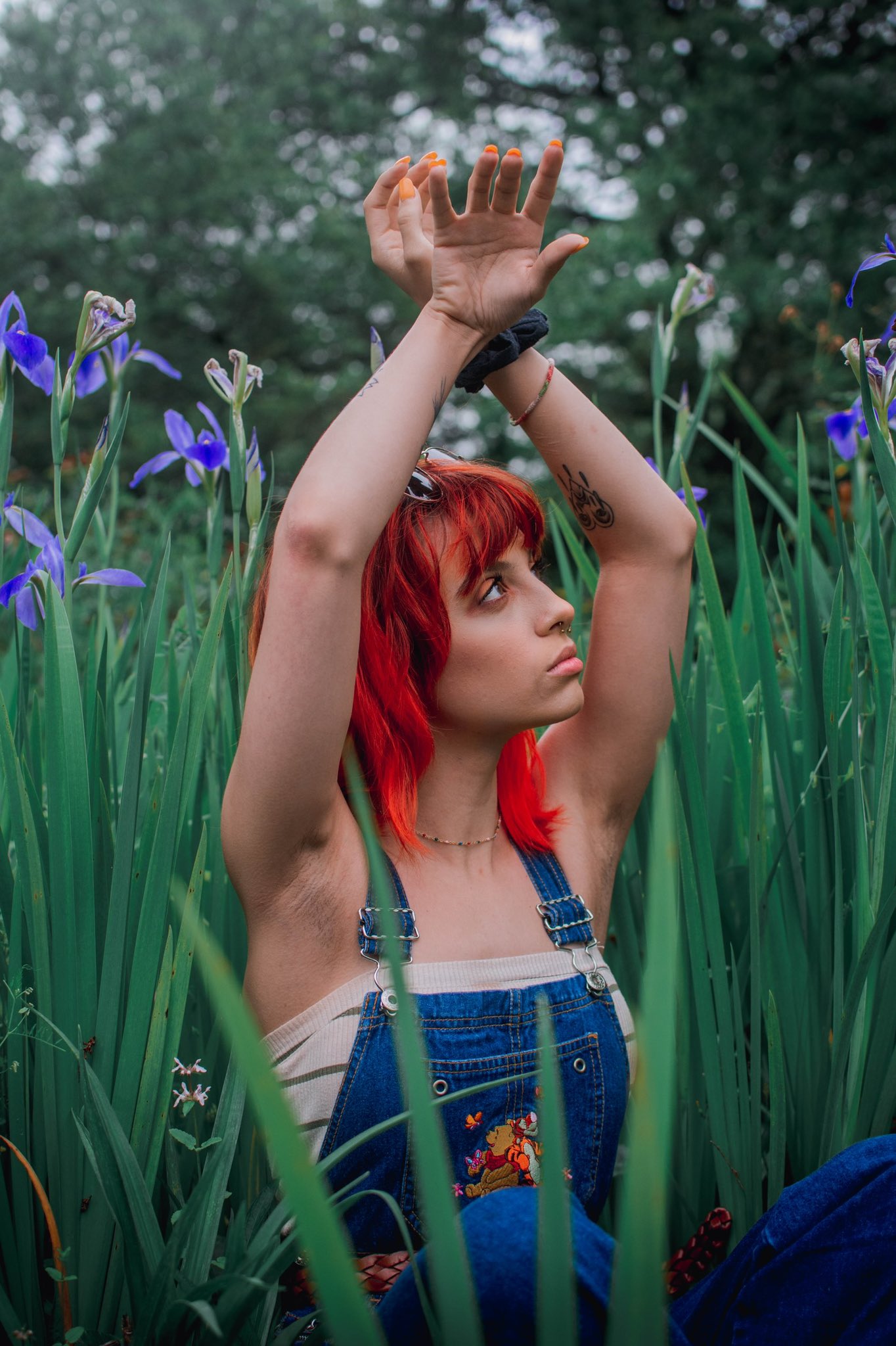 A girl with bright red-orange hair is lifting her arms into the air, her nails painted in a similar shade of orange. She is wearing blue overalls and a white shirt and she is surrounded by extremely tall grass adorned with purple flowers.