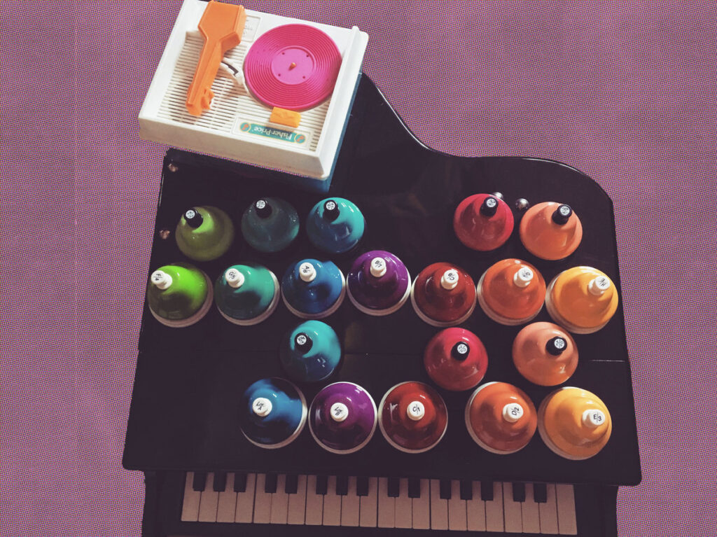 Alexa Dexa’s 37 key Schoenhut baby grand toy piano floats on a purple background. On top of the toy piano sit a very colorful set of 20 chromatic toy desk bells and a toy record player with a custom pink 3D printed toy record which plays a tune Alexa composed for their electroacoustic toy opera, Be a Doll.